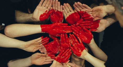 a group of people have pushed their hands together, palm up, and a bright red heart has been painted across all the hands.