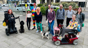 pavement with the street behind them. Most of them are standing, but 1 is seated in a powered wheelchair and 1 in a mobility scooter. One man is dressed in a colourful rainbow suit. They have 3 dogs with them.