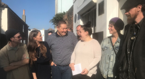 A group of 6 people are stood outside a building on a sunny day. They are all dressed casually and are smiling at each other. The woman in the centre is holding a piece of paper. 