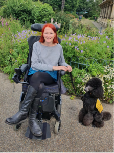 A smiling woman with bobbed red hair sits in a powered wheelchair. Beside her she has a dog on a lead - a large black poodle with a yellow assistance dog jacket on. They are sat in a pretty public garden, surrounded by flowers and greenery. 