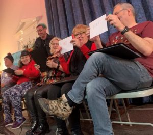 Five people are sat on chairs holding up pieces of cardboard, they are all smiling and laughing. One woman is wearing a soft helmet and a smiling man is stood behind her, supporting her. The people appear to be on a stage with a blue curtain behind them. Everyone looks like they’re having a good time. 