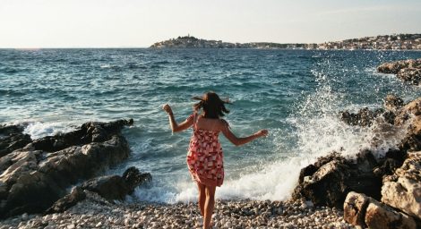 A woman in a red sun dress has her back towards us, she is standing on one foot in front of a wave on a rocky beach. It's a sunny day and we see the spray from the waves on the rocks.