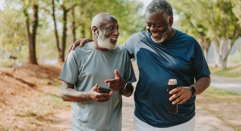 Two older men are walking outside in nature. They are smiling and gesticulating as if they've just shared a joke.